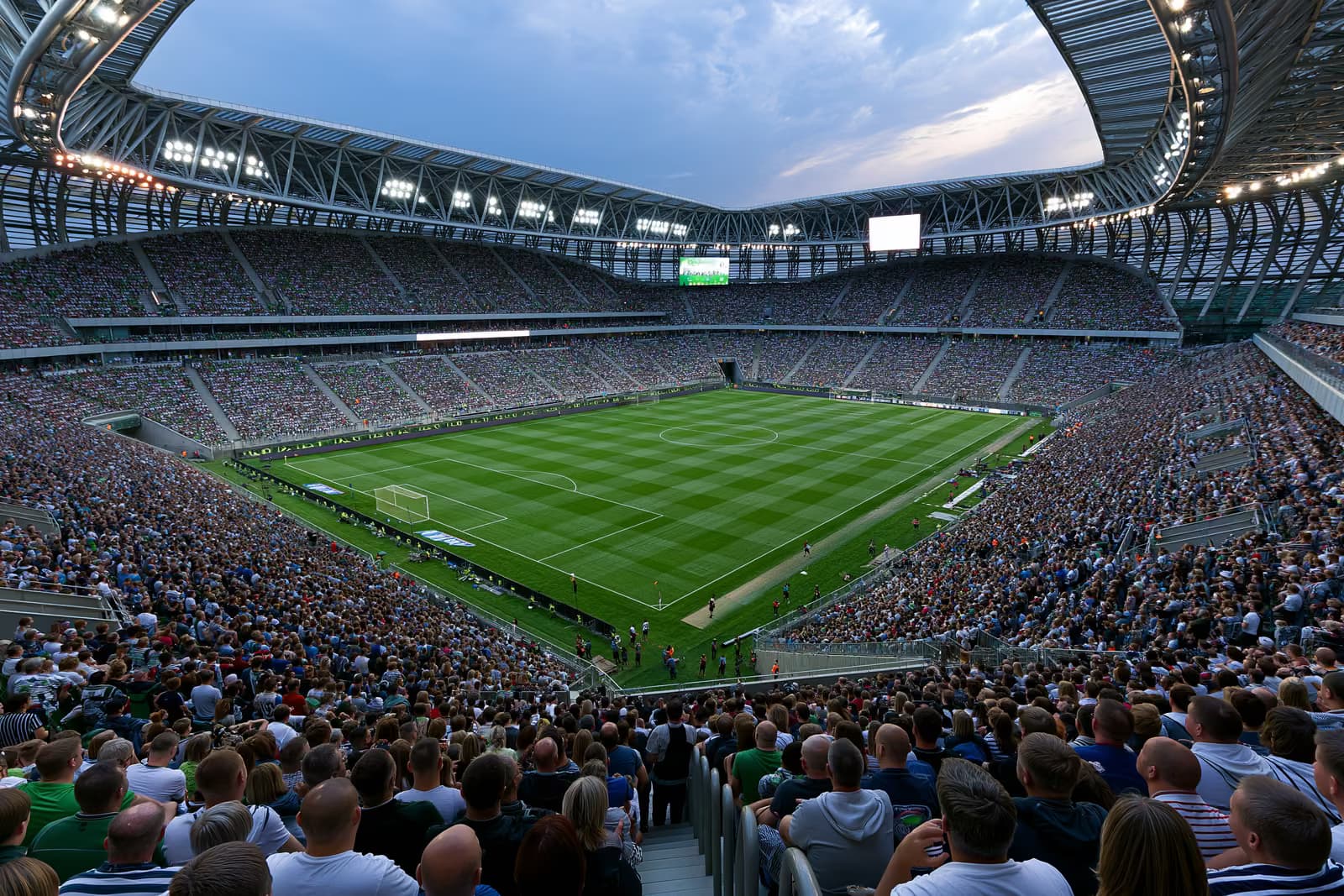 Duży stadion piłkarski wypełniony kibicami, siedzącymi pod sztucznymi światłami, w letni wieczór.