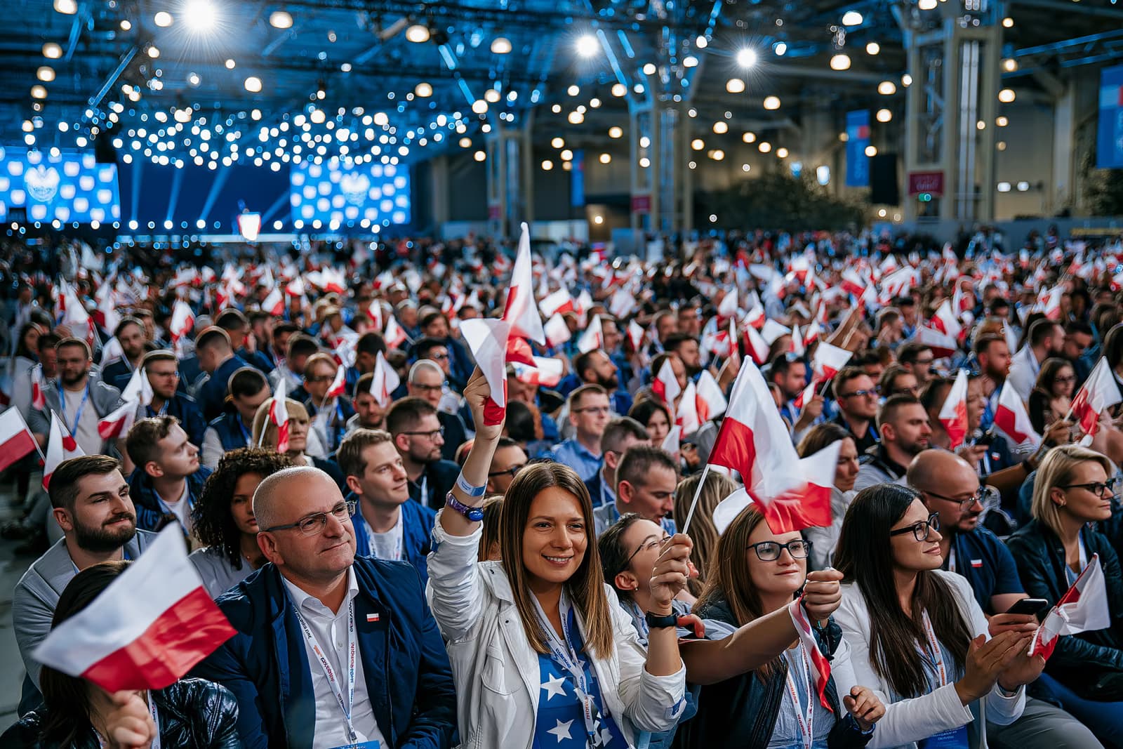 Tłum zgromadzony w pełnej sali wystawowej powiewa małymi flagami w ostrym oświetleniu podczas inauguracji kampanii AfD w Magdeburgu, w oddali widoczna jest scena.