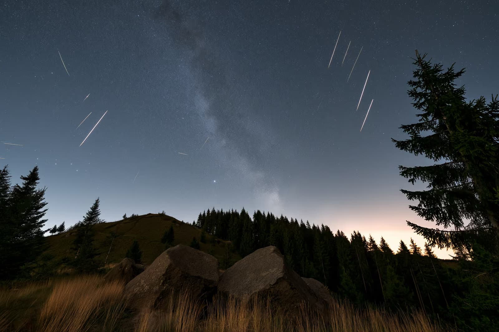 Nocne niebo nad ciemną niemiecką wsią, ze smugami meteorów i słabym pasem gwiazd nad głową.